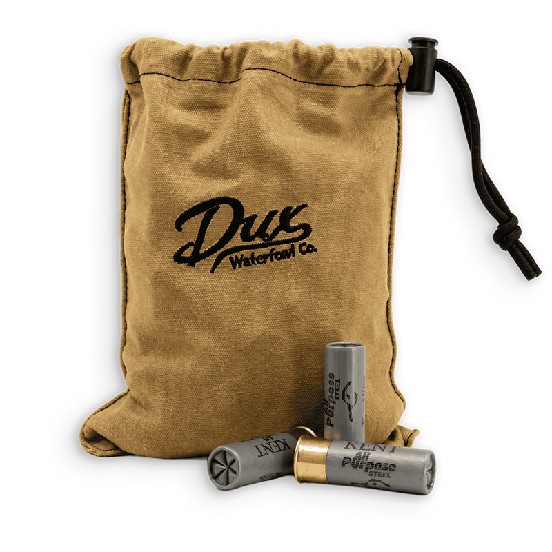 A tan canvas drawstring pouch with black embroidery reading 'Drux Waterfowl Co.' and a black drawcord, shown with two gray shotgun shells in front on a white background.