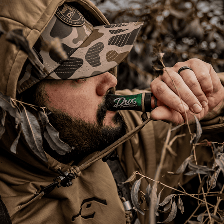 A close-up lifestyle shot of a bearded man in camouflage hunting attire holding a green and black Duz Outdoors shotgun to his mouth, surrounded by dry leaves and branches.