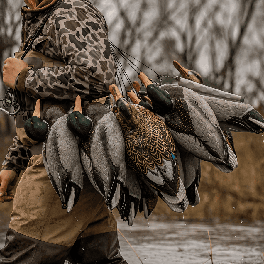 A duck hunter wearing camouflage clothing carries a large bundle of real mallards on their back in an outdoor river setting.