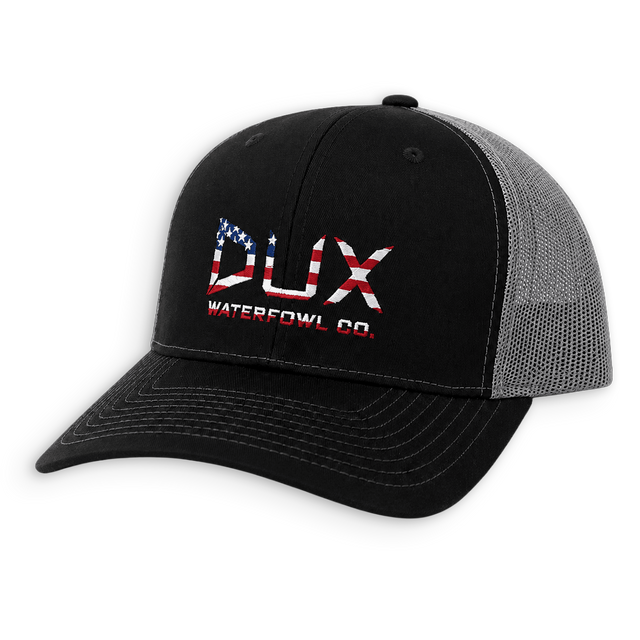 A black trucker-style cap with a gray mesh back, featuring a red, white, and blue embroidered logo on the front that reads 'DUX WATERFOWL CO.' with an American flag motif.