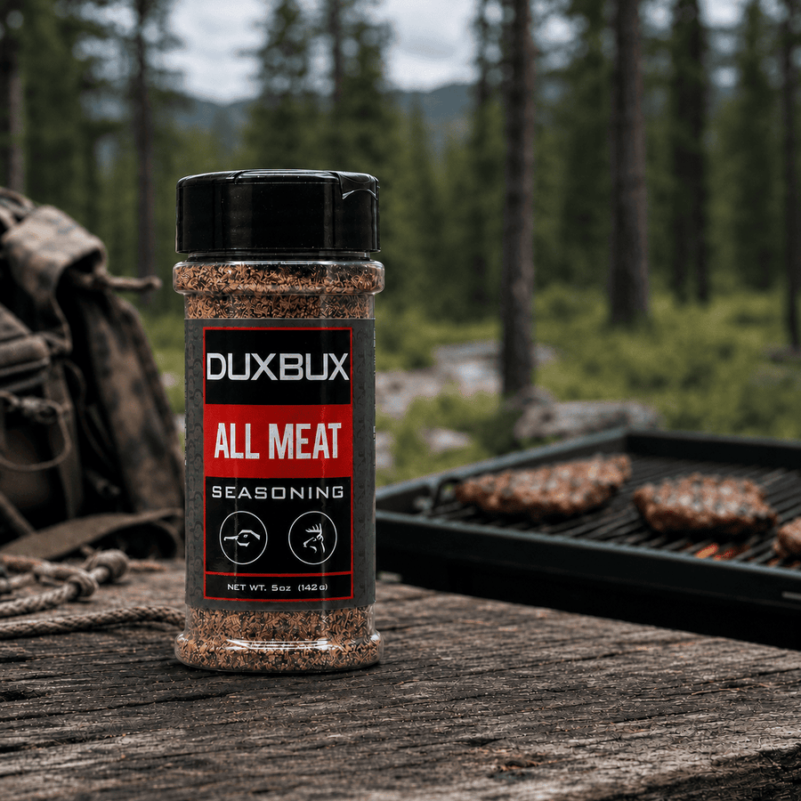 A bottle of DUXBUX All Meat Seasoning stands on a rustic wooden surface with a blurred outdoor forest background and a grill with meat in the distance.