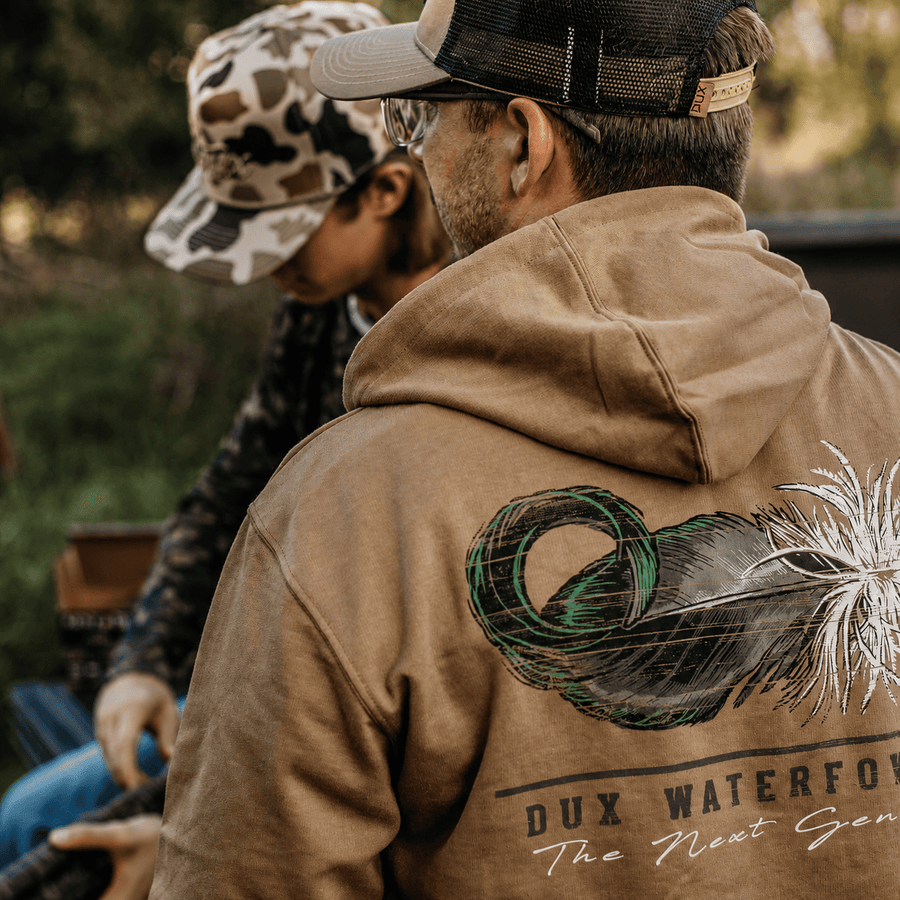 A man wearing a tan Dux Waterfowl hoodie with a large graphic on the back, shown from behind in an outdoor setting. Another person in a camouflage cap is seated in the background.