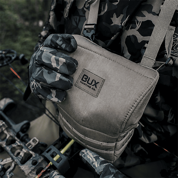 A close-up of a gray canvas crossbody bag worn by a person in camouflage clothing and gloves, with the brand patch reading BUX Hunting Co. visible on the front.