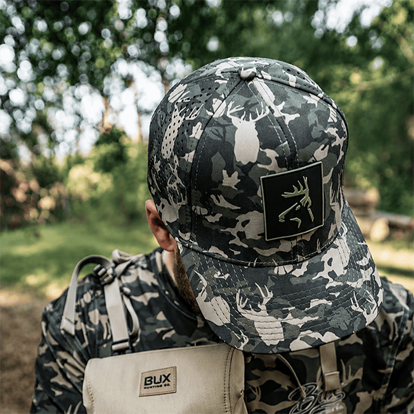 A man outdoors wearing a dark gray and black camouflage baseball cap with a black square patch featuring a deer head logo, along with a matching camouflage shirt and a beige BUX bag.
