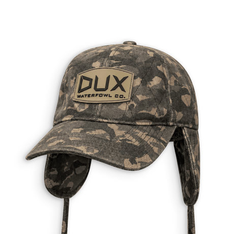 A Dux Waterfowl Co. camouflage winter cap with ear flaps and chin straps, shown on a plain white background.