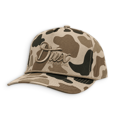 A baseball cap in a beige, brown, and black camouflage pattern, featuring a prominent metallic bronze 'Dux Waterfowl' logo on the front panel.