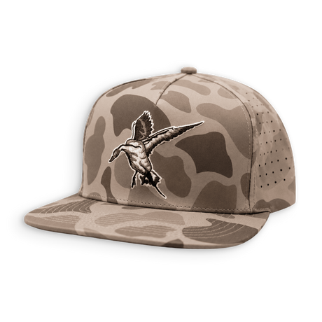 A flat-brim snapback cap in a desert camouflage pattern, featuring a detailed embroidered duck hunting duck on the front panel. The cap has a tan and brown camo pattern with a matching flat brim and perforated side panels.