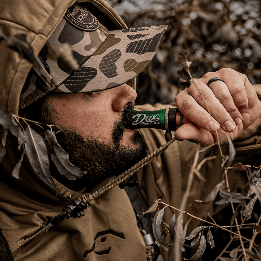 A close-up lifestyle shot of a bearded man in camouflage hunting attire holding a green and black Duz Outdoors shotgun to his mouth, surrounded by dry leaves and branches.