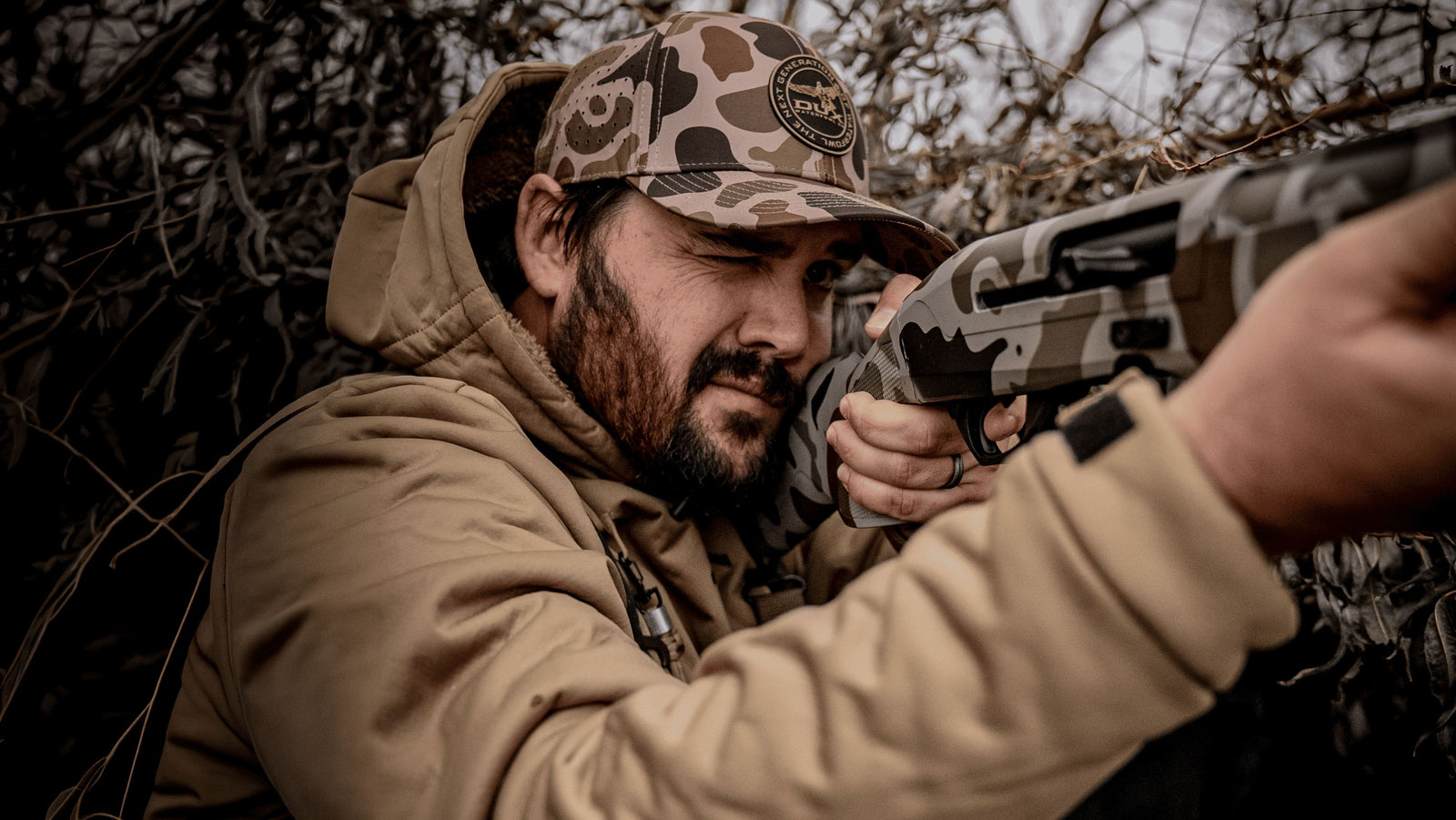 Andrew Flair in a tan hunting jacket and camouflage cap aims a camouflage-patterned shotgun while sitting among branches in a forest setting.