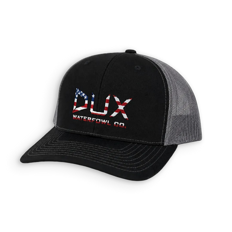 A black and gray trucker hat with a curved brim, featuring a front panel logo that reads 'DUX WATERFOWL CO.' in red, white, and blue with an American flag pattern.