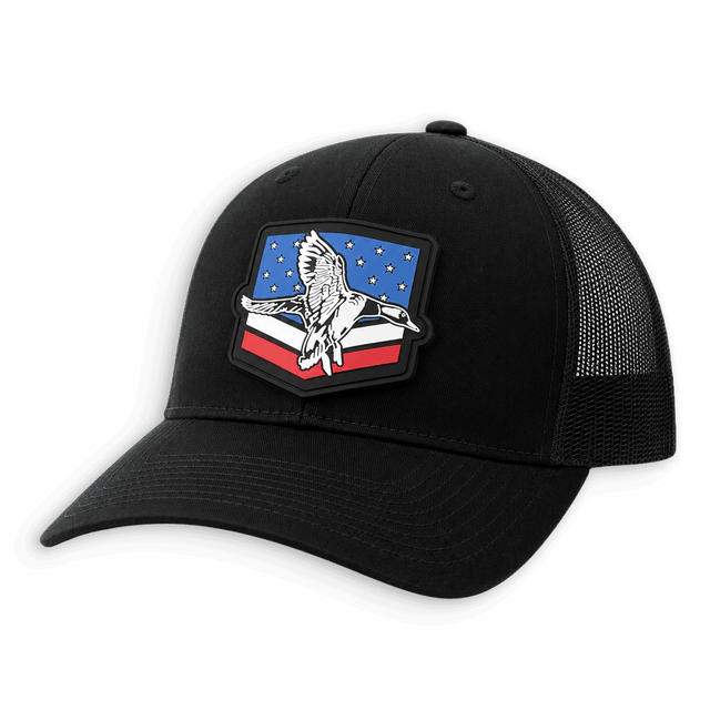 A black trucker hat with a curved brim and black mesh back panels, featuring a prominent shield-shaped patch on the front with a white eagle over a red, white, and blue background.